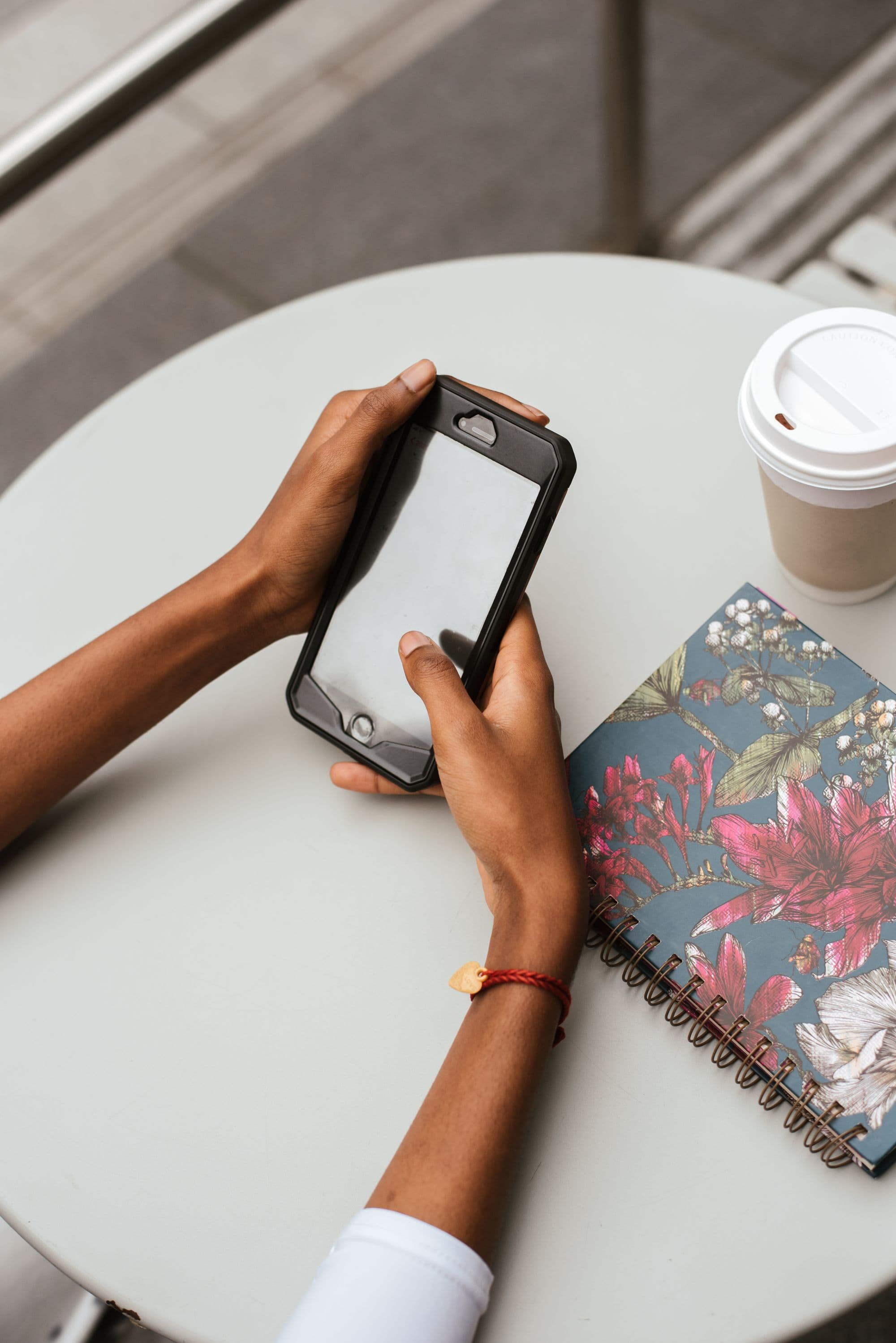 Hands using a smartphone beside a floral notebook and takeaway coffee